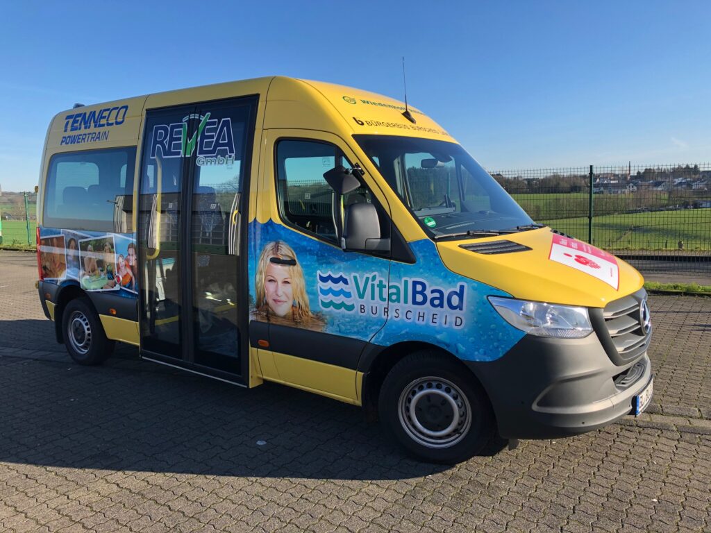 A Mercedes-Benz Sprinter 8-seater minibus is shown. It is yellow in colour and displays five large format  adverts for local businesses. The bus has a large door at the centre that swings out and to the sides and allows for a lower entry point into the vehicle for better accessibility.